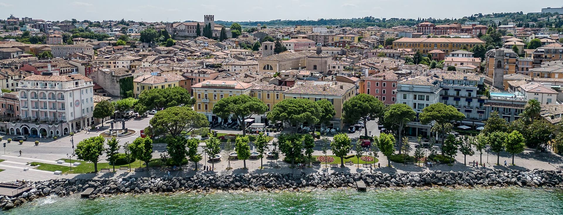 Aerial view of a coastal Italian town with historic buildings, tree-lined promenade, and rocky shoreline along the green sea.