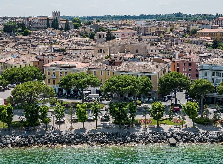 Luftansicht einer italienischen Küstenstadt mit historischen Gebäuden, baumumsäumter Promenade und felsiger Küstenlinie entlang des grünen Meeres.