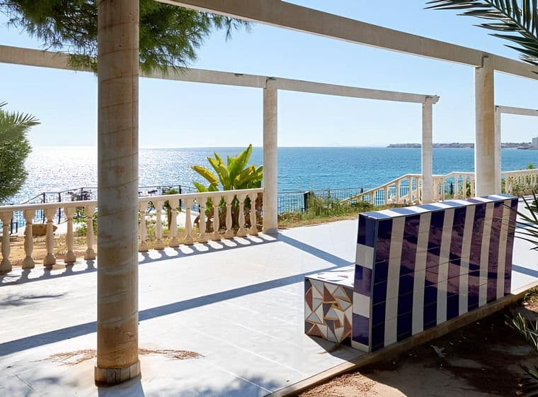 Banca fixa buida al passeig de Punta Prima amb vista al mar i palmeres.