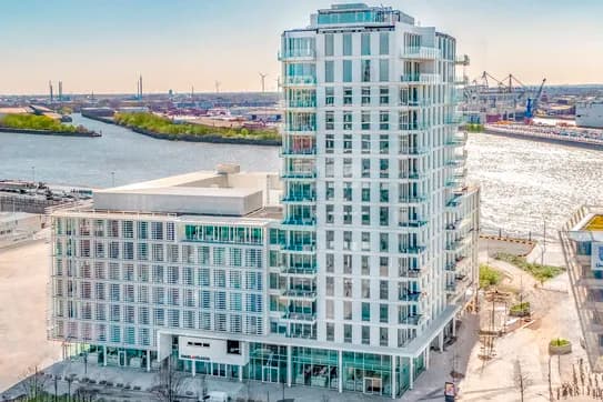 Modernes, glasverkleidetes Wohnhochhaus am Flussufer, mit einem in der Ferne liegenden Hafen, Kränen und Windrädern unter strahlend blauem Himmel.