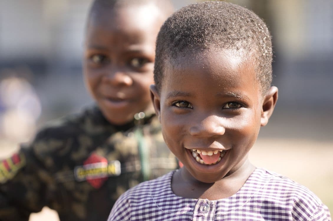 Two smiling children, with one in the foreground wearing a checkered shirt and the other in a camo jacket, outdoors in daylight.