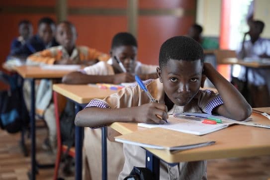 A student in a classroom focuses on writing in a notebook. Other students are seated at desks in the background.