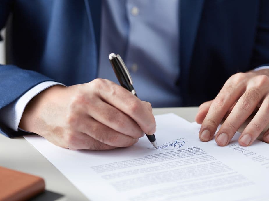 Man in a blue suit signs a contract document on a desk with a fountain pen.