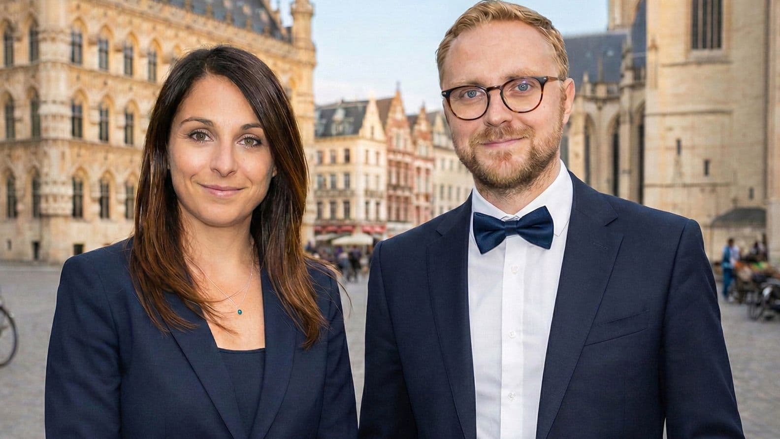 A man and woman in formal attire stand smiling outside historic European buildings on a clear day.