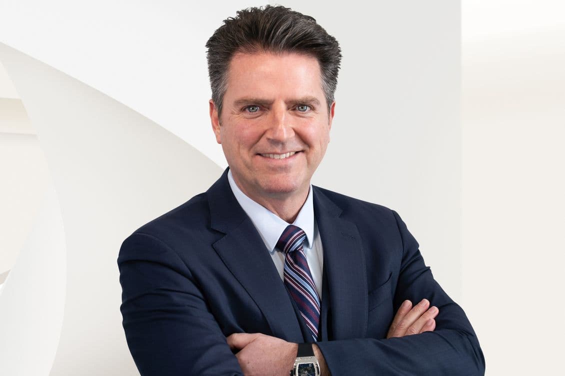 Portrait d'un agent immobilier : Homme souriant en costume bleu marine, cravate rayée et montre, les bras croisés sur un fond blanc.