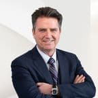 Real estate agent portrait: Smiling man in a navy suit, striped tie, and watch, arms crossed against a white background.