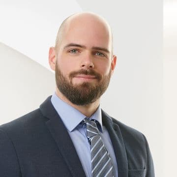 A man with a beard and shaved head in a suit and tie smiles slightly against a light, abstract background.