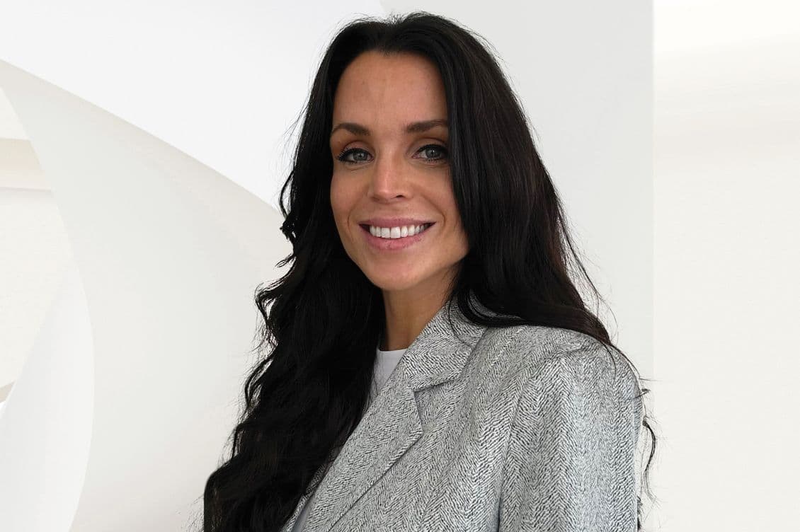 A smiling woman with long dark hair, wearing a gray blazer, stands against a white background.