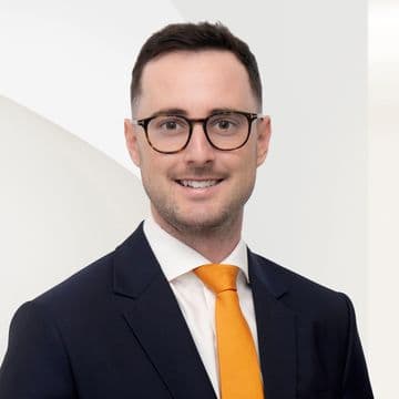 A man in a dark suit and orange tie, wearing glasses, smiling against a white, modern background.
