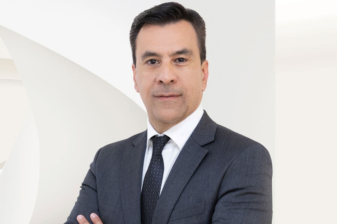 Ένας άνδρας με σκούρο κοστούμι, λευκό πουκάμισο και σκούρα γραβάτα στέκεται με σταυρωμένα χέρια μπροστά σε ένα λευκό, μοντέρνο φόντο, κοιτάζοντας απευθείας την κάμερα με ουδέτερη έκφραση.