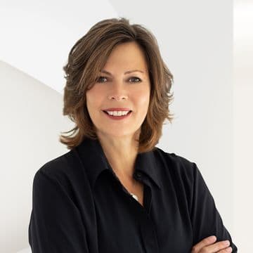 Smiling woman with short brown hair and a black shirt, standing in front of a white geometric background.