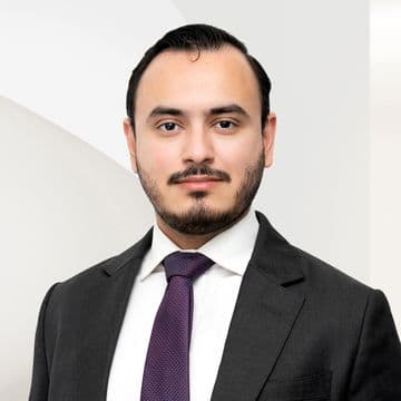 Businessperson in a dark suit, white shirt, and maroon tie, posing against a minimalist, light-colored background.