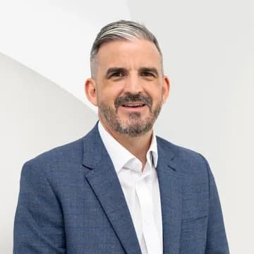 Portrait of a middle-aged man with salt-and-pepper hair and beard, wearing a blue suit jacket and white shirt against a pale geometric backdrop.