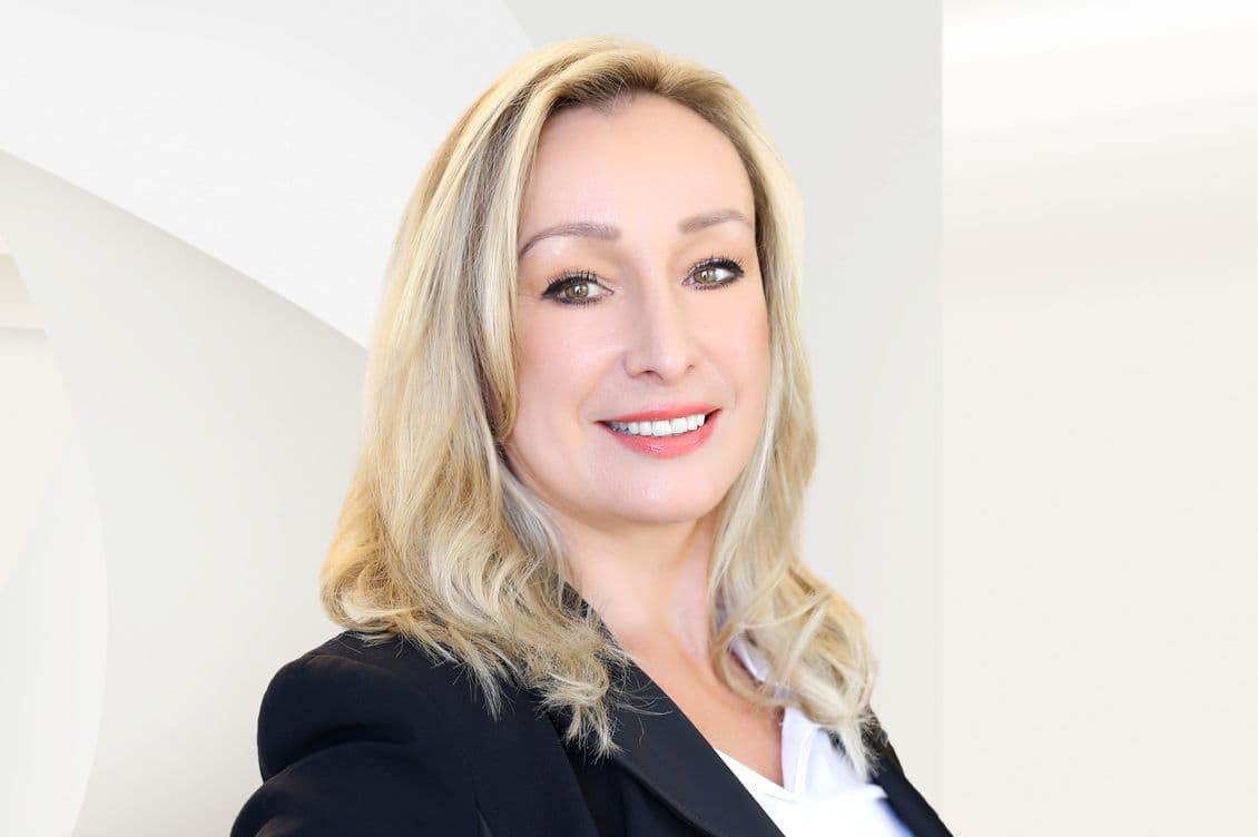 Smiling woman with long blonde hair in a black blazer and white shirt, standing against a light, abstract background.