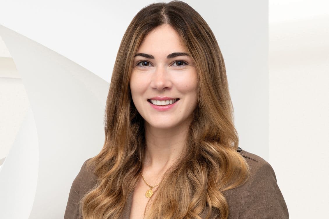 Smiling young woman headshot with long wavy brown hair, brown blazer and gold pendant necklace on a light background.