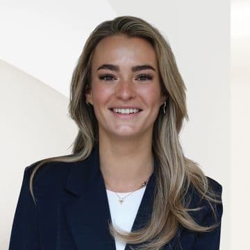 Headshot of a smiling woman with blonde hair, wearing a navy blazer and gold jewelry, against a white background.