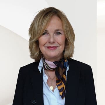 Headshot of a blonde woman in a black blazer, white shirt, and patterned scarf. She wears a gold necklace and smiles against a white background.