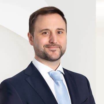 A man in a dark suit with a light blue tie stands against a white background with a neutral expression.