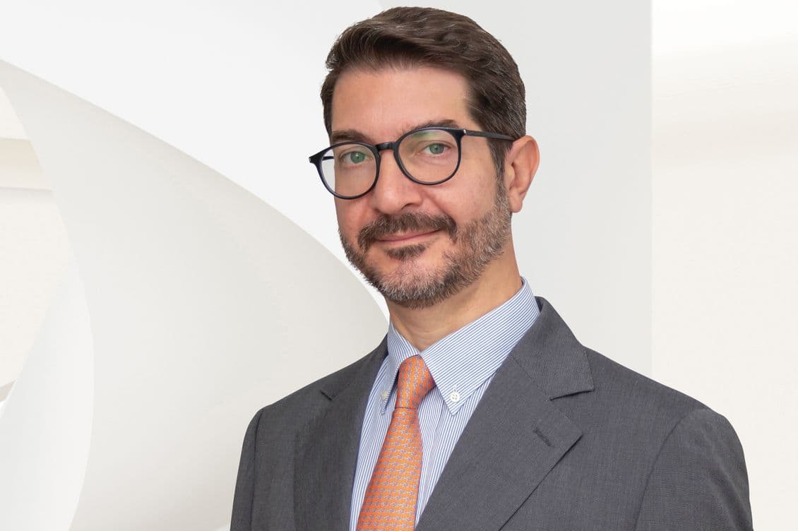 A man with glasses, a beard, and a gray suit smiles against a white background.