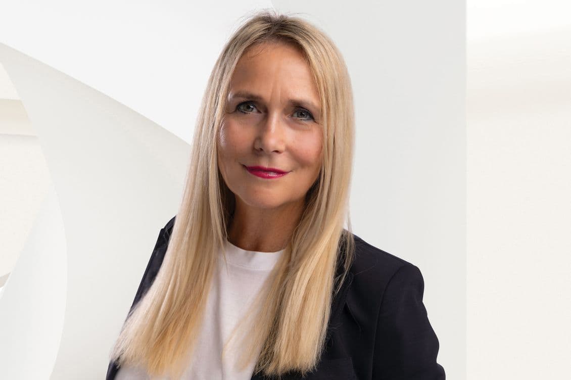 Portrait of a professional woman with long straight blonde hair wearing a black blazer over a white top. She has bright pink lipstick and is smiling slightly at the camera. The background is minimalist with white walls, creating a clean, contemporary aesthetic for this professional headshot. The image has a polished, corporate feel suitable for a business profile or company website.