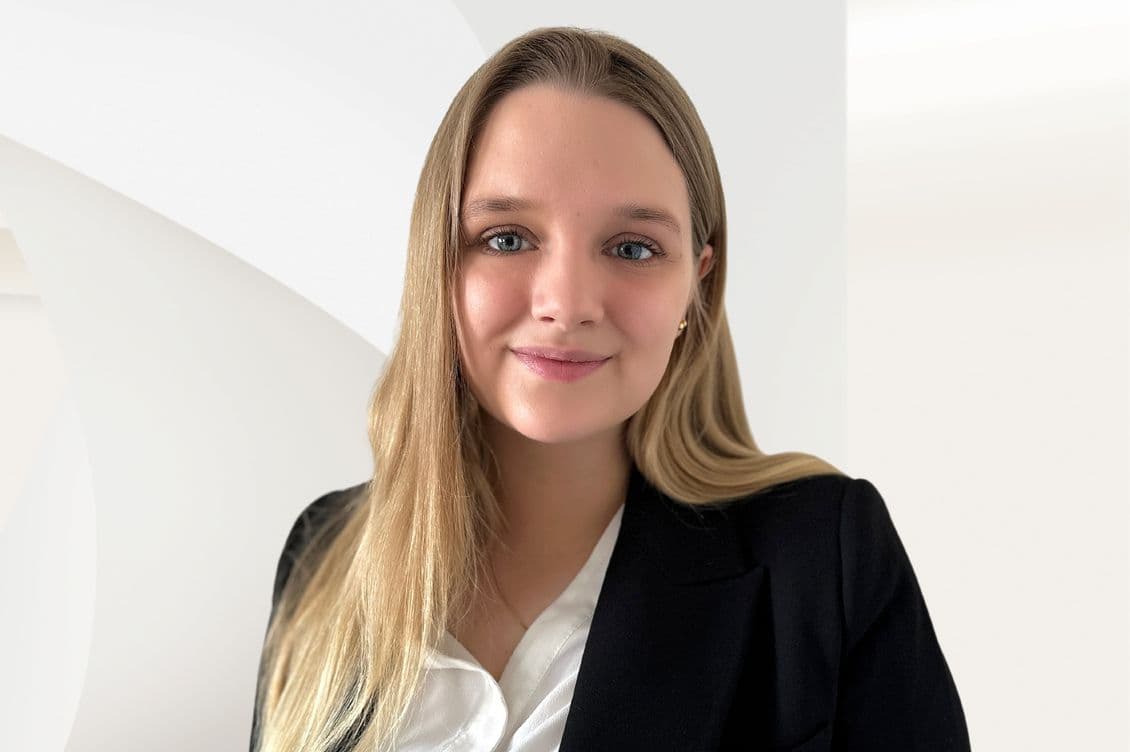 Frau mit langen blonden Haaren in einem schwarzen Blazer und weißem Hemd, lächelnd vor einem hellen, abstrakten Hintergrund.