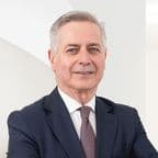 A distinguished senior businessman with gray hair stands confidently against a white background. He wears a dark blue suit with a patterned tie, exuding professionalism and experience. His warm smile suggests approachability, making him appear as a knowledgeable consultant on property prices in Rome. The image conveys trust and expertise in the Roman real estate market.