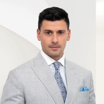 Luca Cappalonga in a light gray suit with a blue tie and pocket square, standing against a white background with geometric shapes.