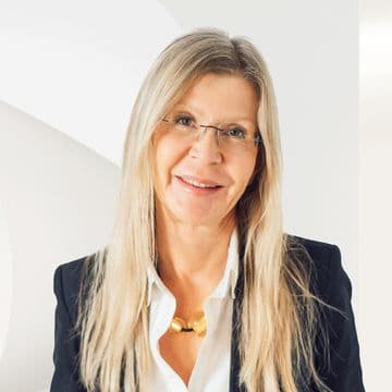 Smiling woman with long blonde hair, wearing glasses and a black blazer, in front of a modern, white abstract background.