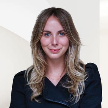 Real estate agent headshot: Smiling woman with blonde hair and blue eyes, wearing a black jacket against a light background.