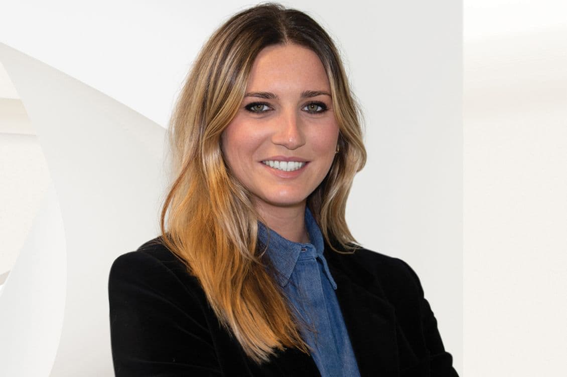 A smiling woman with long blonde hair, wearing a blue denim shirt and black blazer, poses against a white background.
