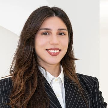 Woman with long brown hair smiling, wearing a black pinstripe blazer and white shirt, standing in a bright, minimalist indoor setting.
