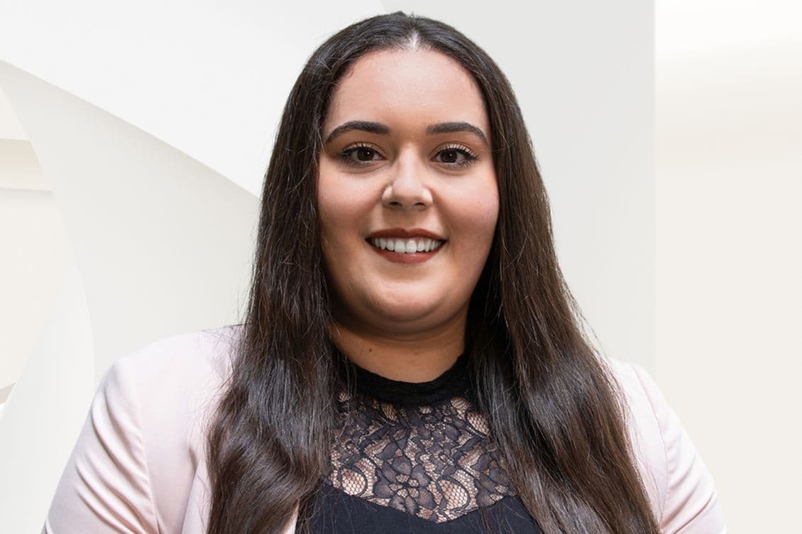 Headshot of a smiling woman with long brown hair, wearing a pink blazer and black lace top, against a light background.