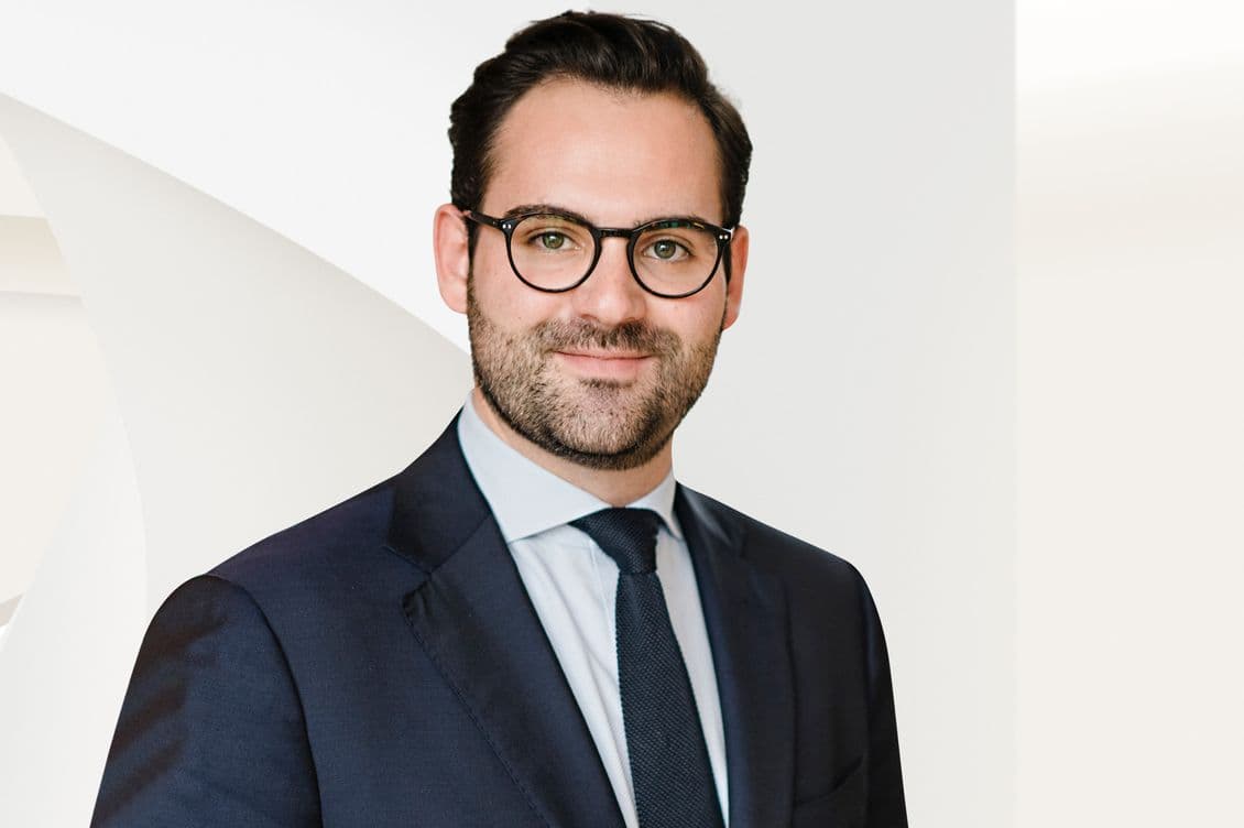 Ein Mann in einem dunklen Anzug und mit Krawatte, der eine Brille trägt, steht vor einem hellen Hintergrund und lächelt leicht.