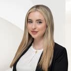 Headshot of a young woman with long blonde hair, wearing a black blazer and gold necklace, against a white background.