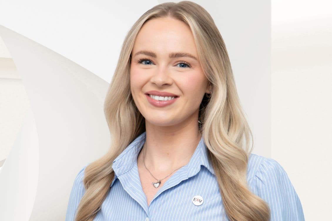 Smiling blonde woman in light blue striped shirt with heart necklace and E&V pin against a pale geometric background.