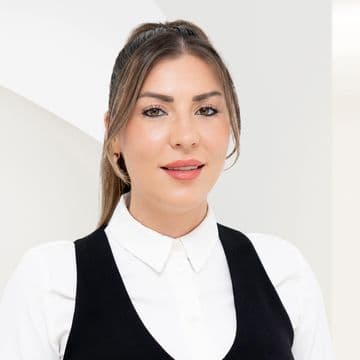 A woman with long hair tied back, wearing a white blouse and black vest, stands against a white background, smiling slightly.