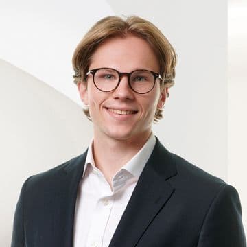 Eine lächelnde Person mit Brille und braunen Haaren, die einen dunklen Blazer und ein weißes Hemd trägt, vor einem hellen, abstrakten Hintergrund