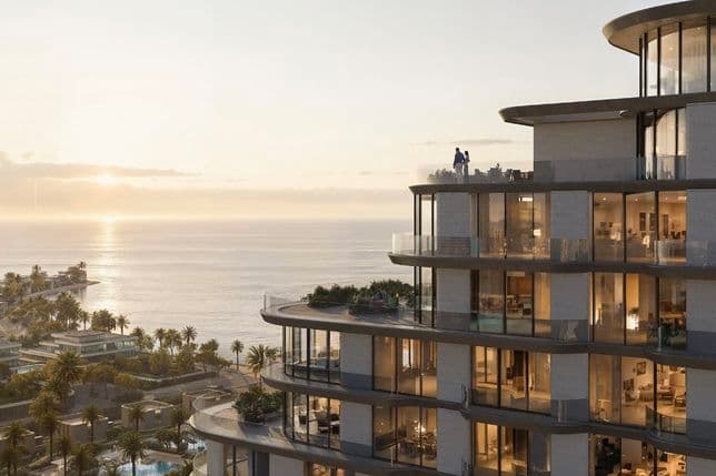 Modern high-rise building with glass balconies overlooking the ocean at sunset, surrounded by palm trees and coastal scenery.