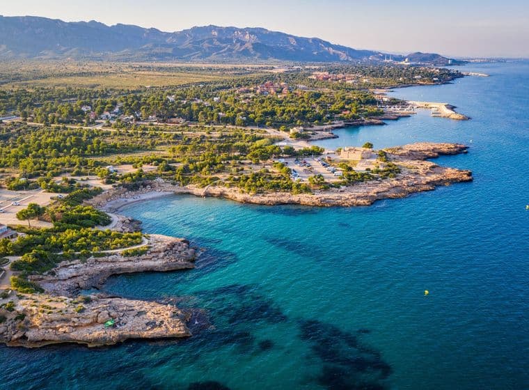 "Es veu la costa, amb algunes platges, alguns edificis, muntanyes al fons i el mar a la dreta de la fotografia."