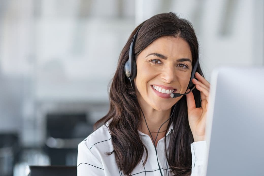 A smiling woman with dark hair wears a headset and white shirt in an office setting.
