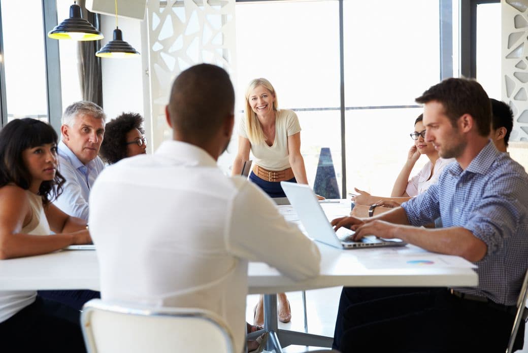 A diverse group of people sit around a white table, with a blonde woman standing at the head, smiling.
