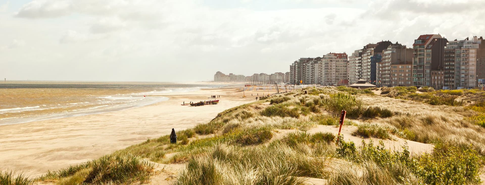 Sandy beach and wind-swept dunes in foreground, small rescue boat and people near shoreline, beachfront apartment buildings under a cloudy sky.