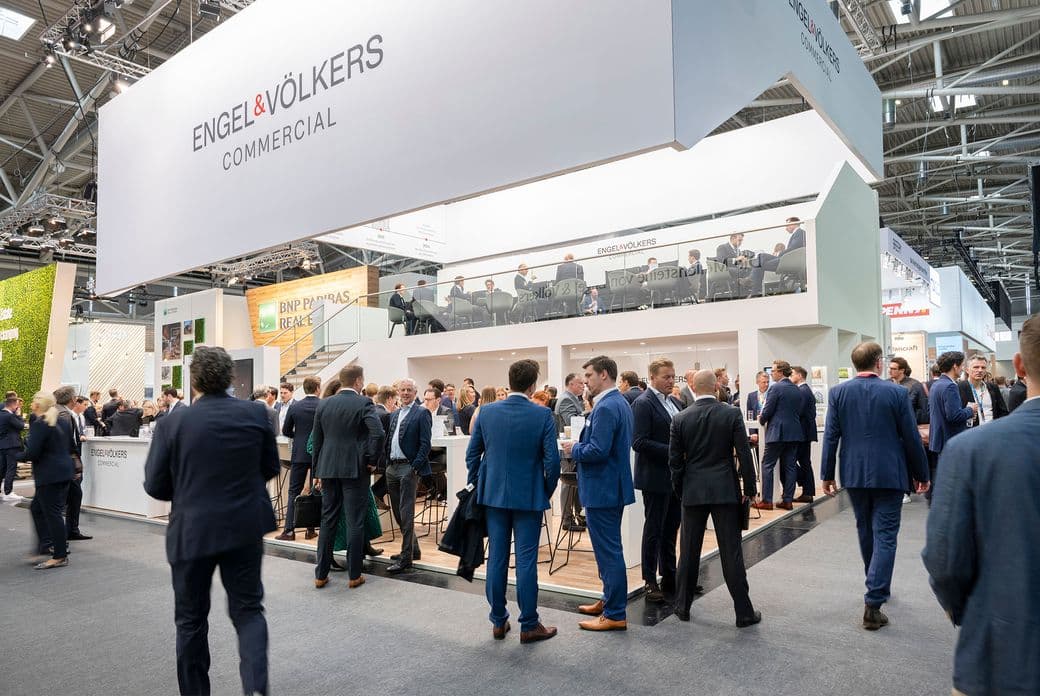 Modern white exhibition booth of Engel & Völkers Commercial with large company branding, surrounded by business professionals in suits networking at a real estate trade show