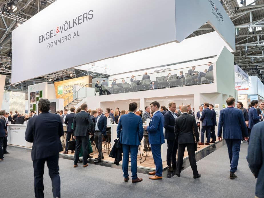 Modern white exhibition booth of Engel & Völkers Commercial with large company branding, surrounded by business professionals in suits networking at a real estate trade show