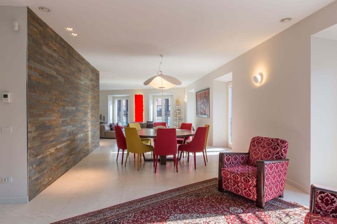 Moderner Essbereich mit Holztisch, bunten Stühlen und gemusterter Wand. Rote Sessel und ein Teppich verleihen dem offenen Raum Wärme.