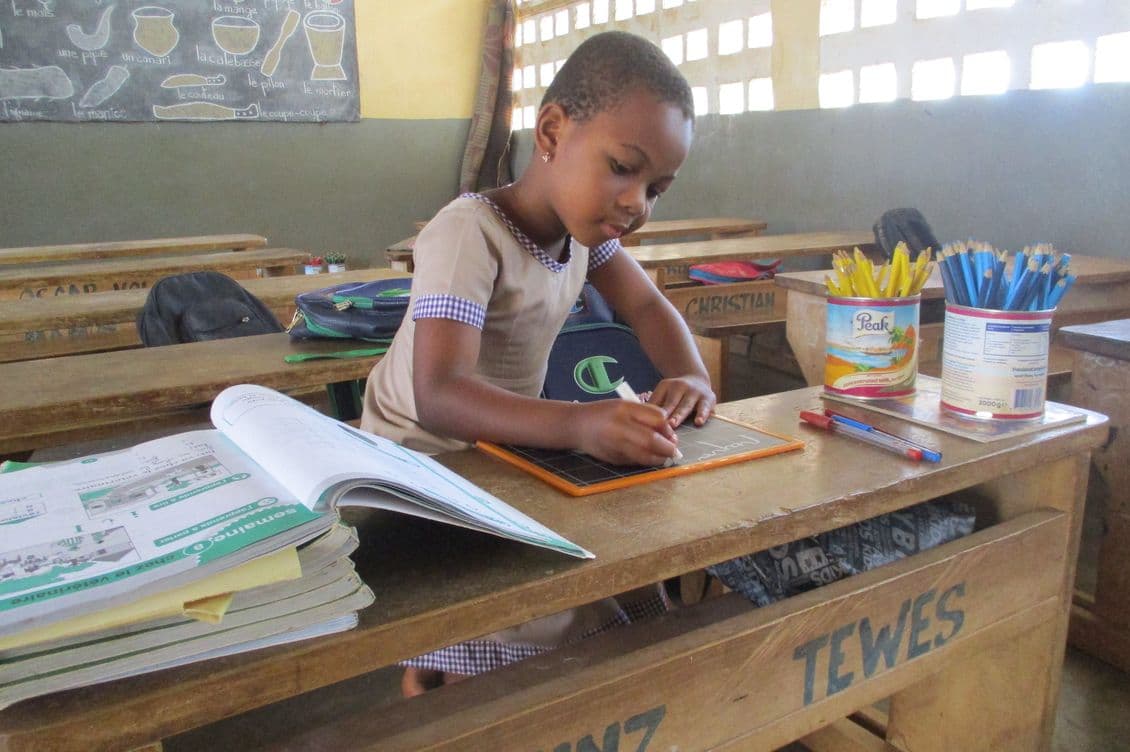 A young child writes on a chalkboard while seated at a wooden desk in a classroom, surrounded by books and pencils.