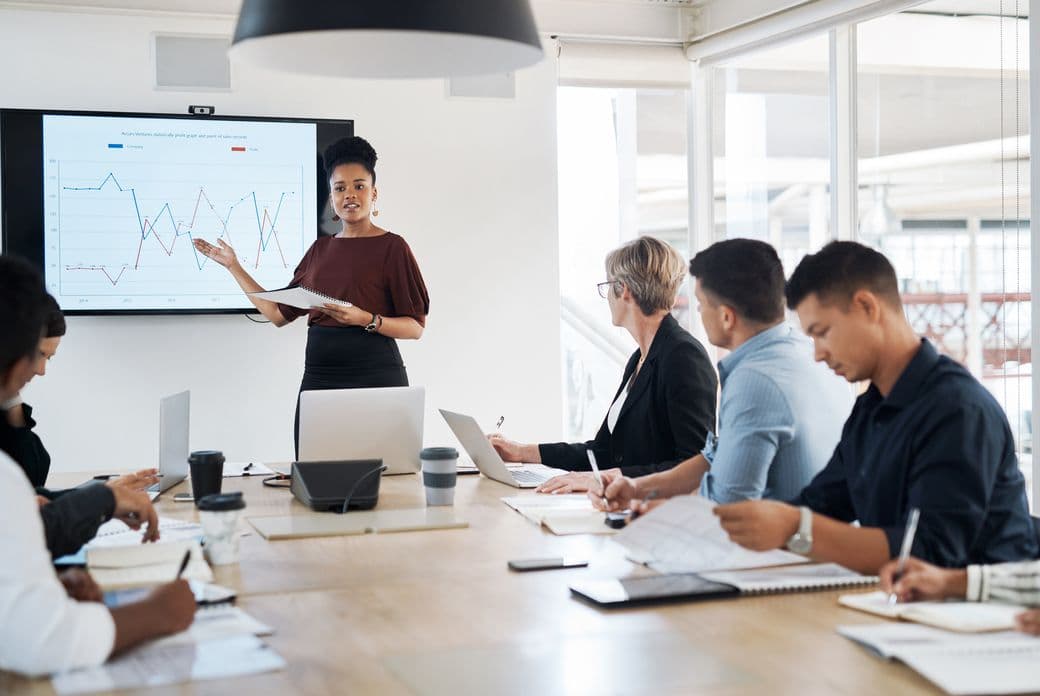 A woman presents a graph to colleagues at a conference table in a bright office.