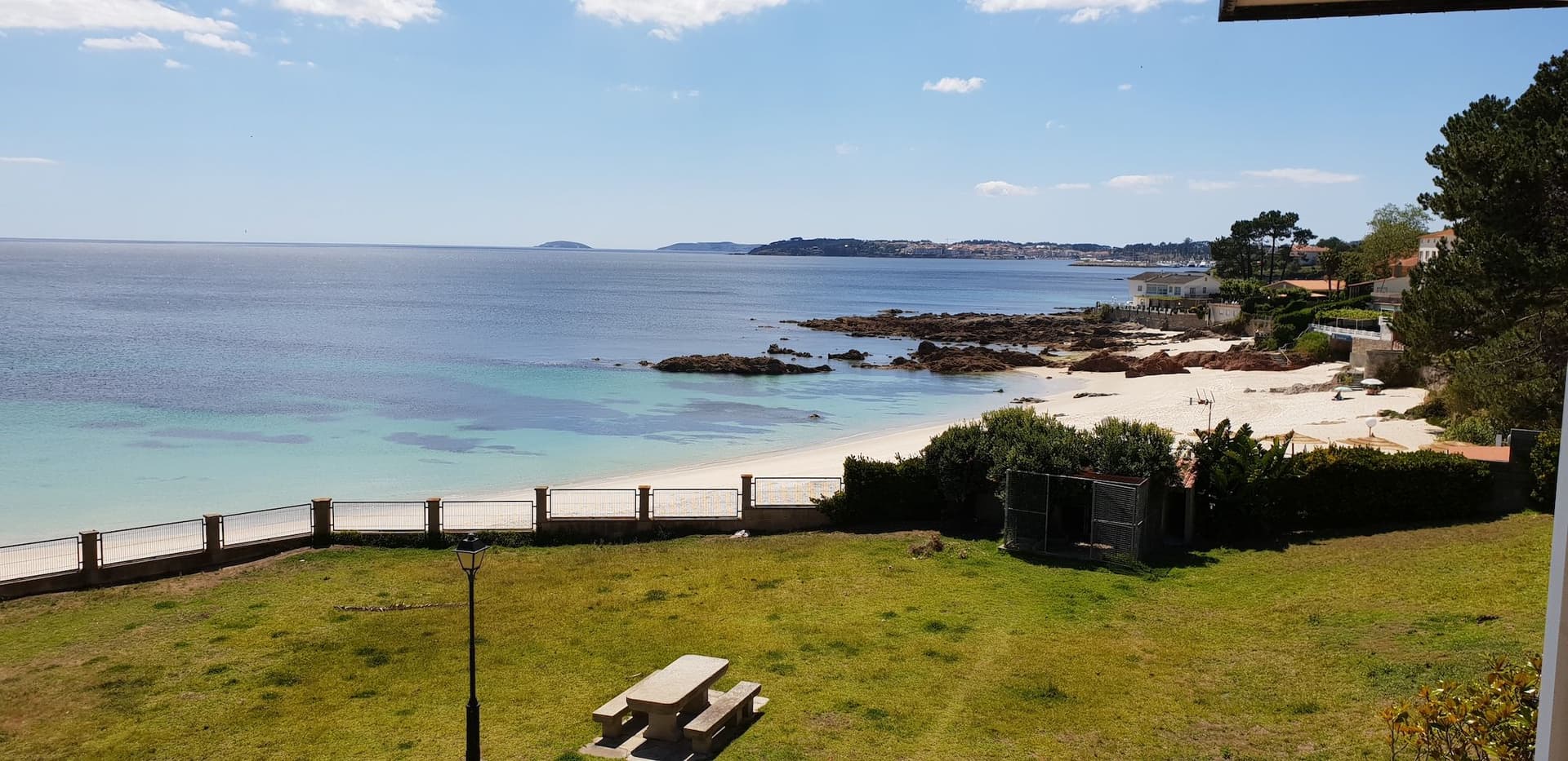 Vista de la platja d'Areas amb Sanxenxo al fons. Es veu un parc amb una taula, la platja al mig i Sanxenxo a l'horitzó.