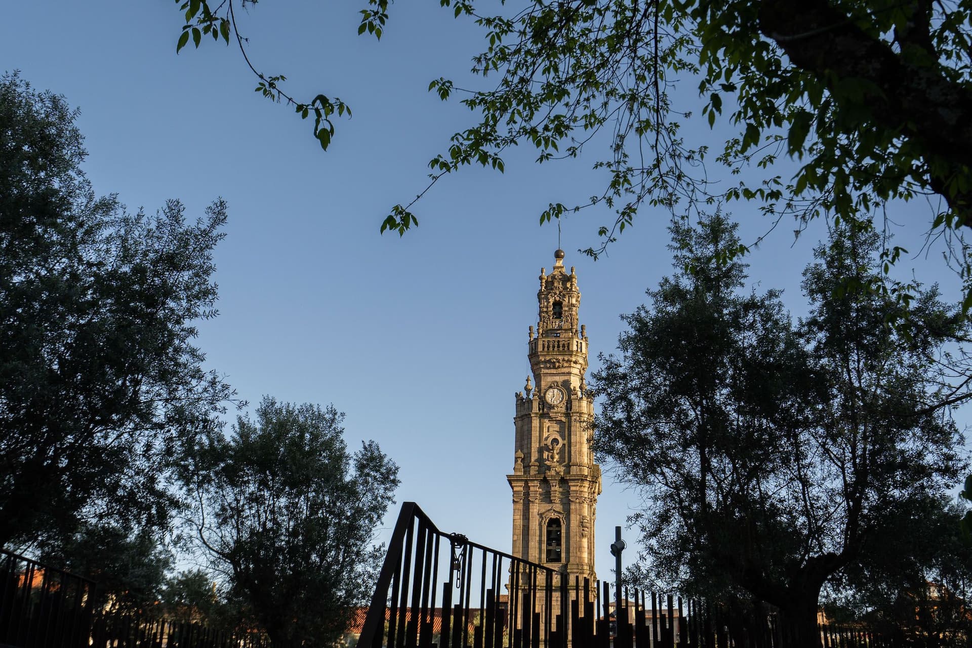 Tall stone tower with a clock, framed by trees and a wrought iron fence, under a clear blue sky.