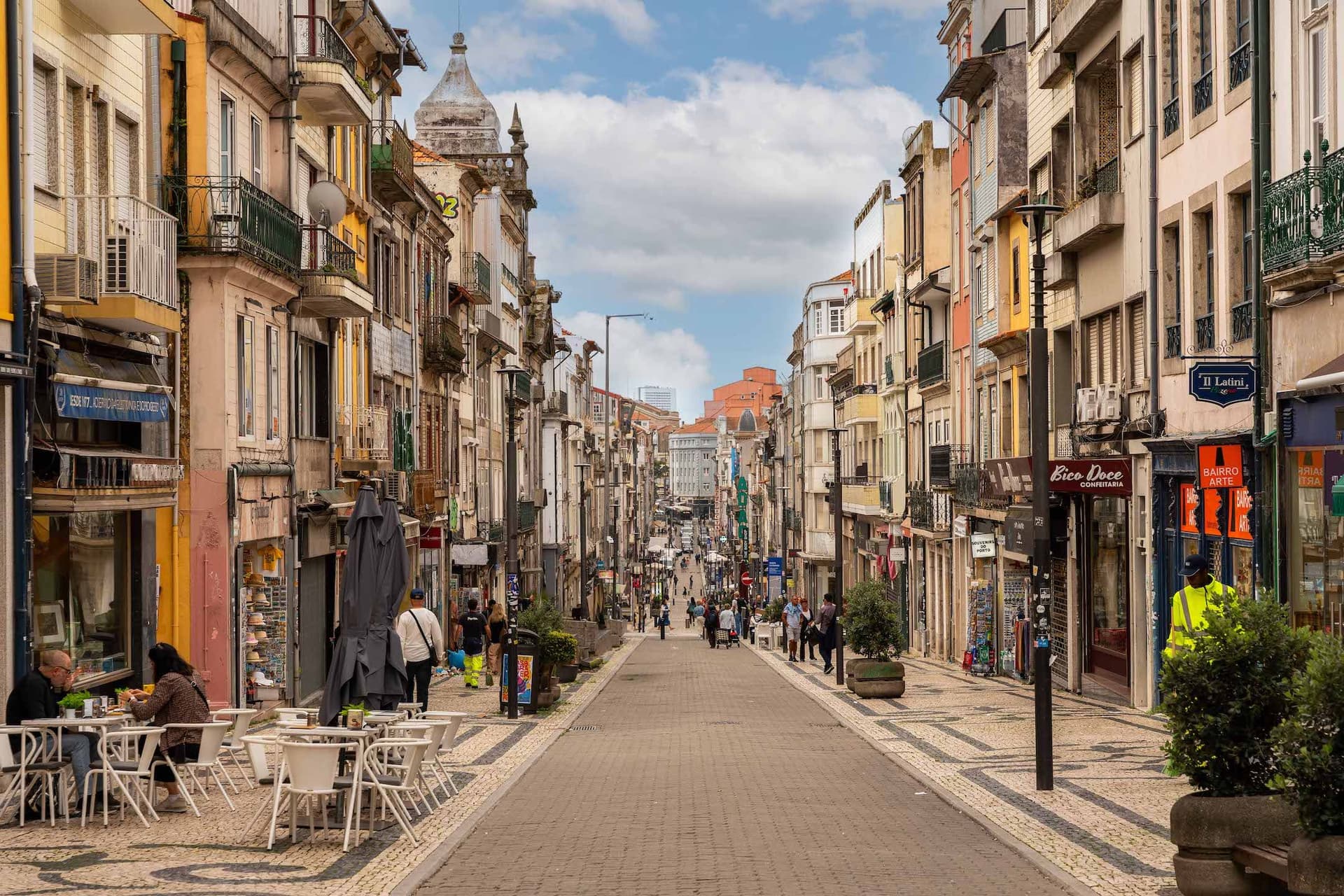 A bustling European street with colorful buildings, outdoor cafes, and pedestrians, under a partly cloudy sky.